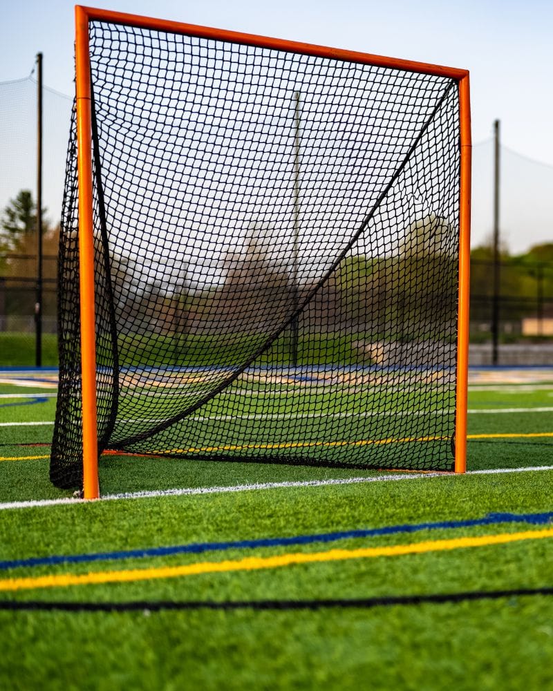 picture of a lacrosse net sitting on an artificial grass lacrosse field in Arizona. The artificial grass is green, and the field markings are black, yellow, blue and white. 