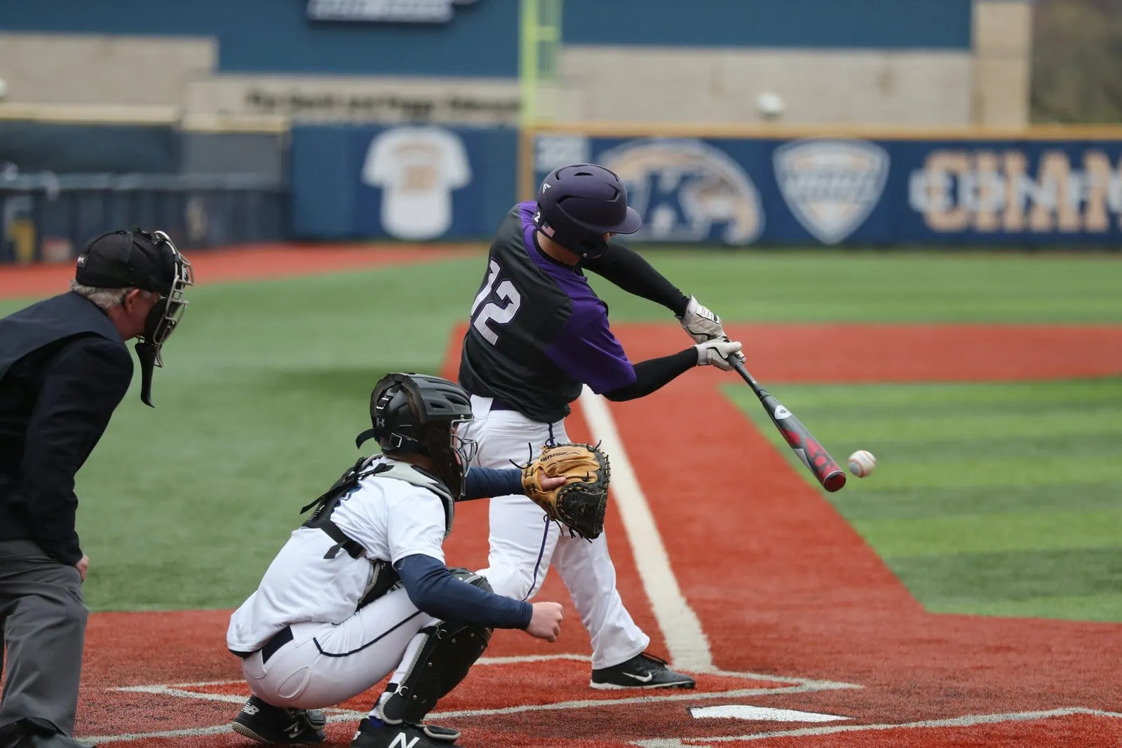 Picture of a baseball player hitting a baseball. Catcher and umpire behind him. Artificial grass baseball field in the background, with red artificial turf on the base paths, and green artificial turf installed on the infield and outfield